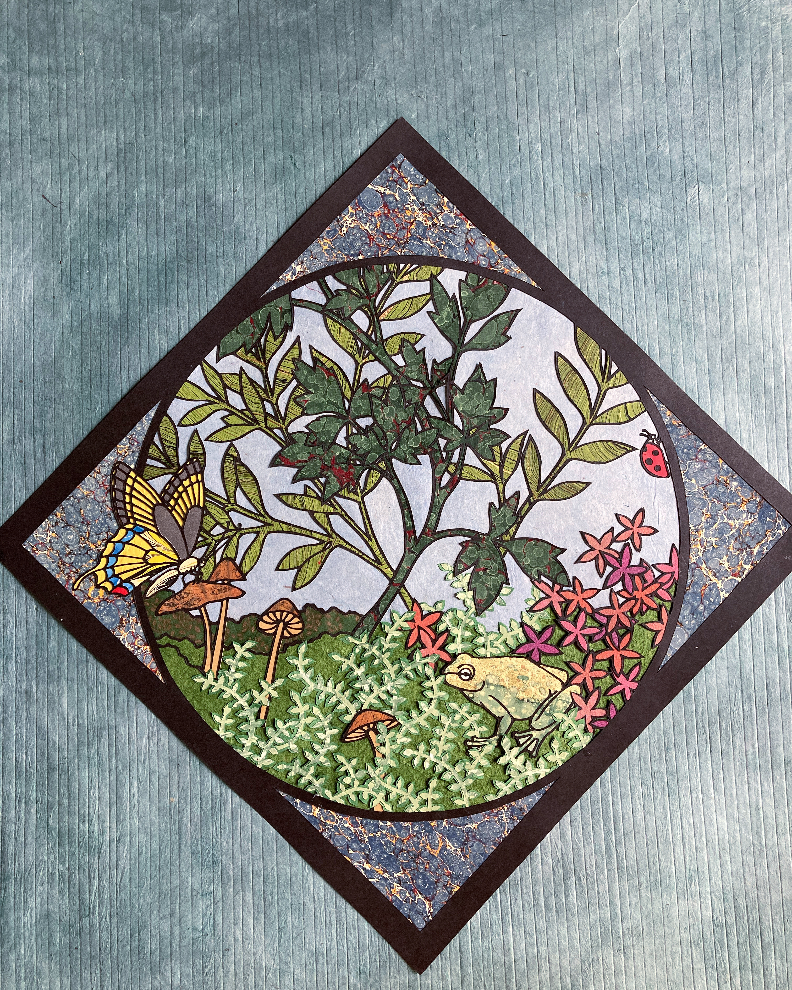 Paper cut artwork featuring a frog, butterfly, mushrooms, and flowers in a circular composition